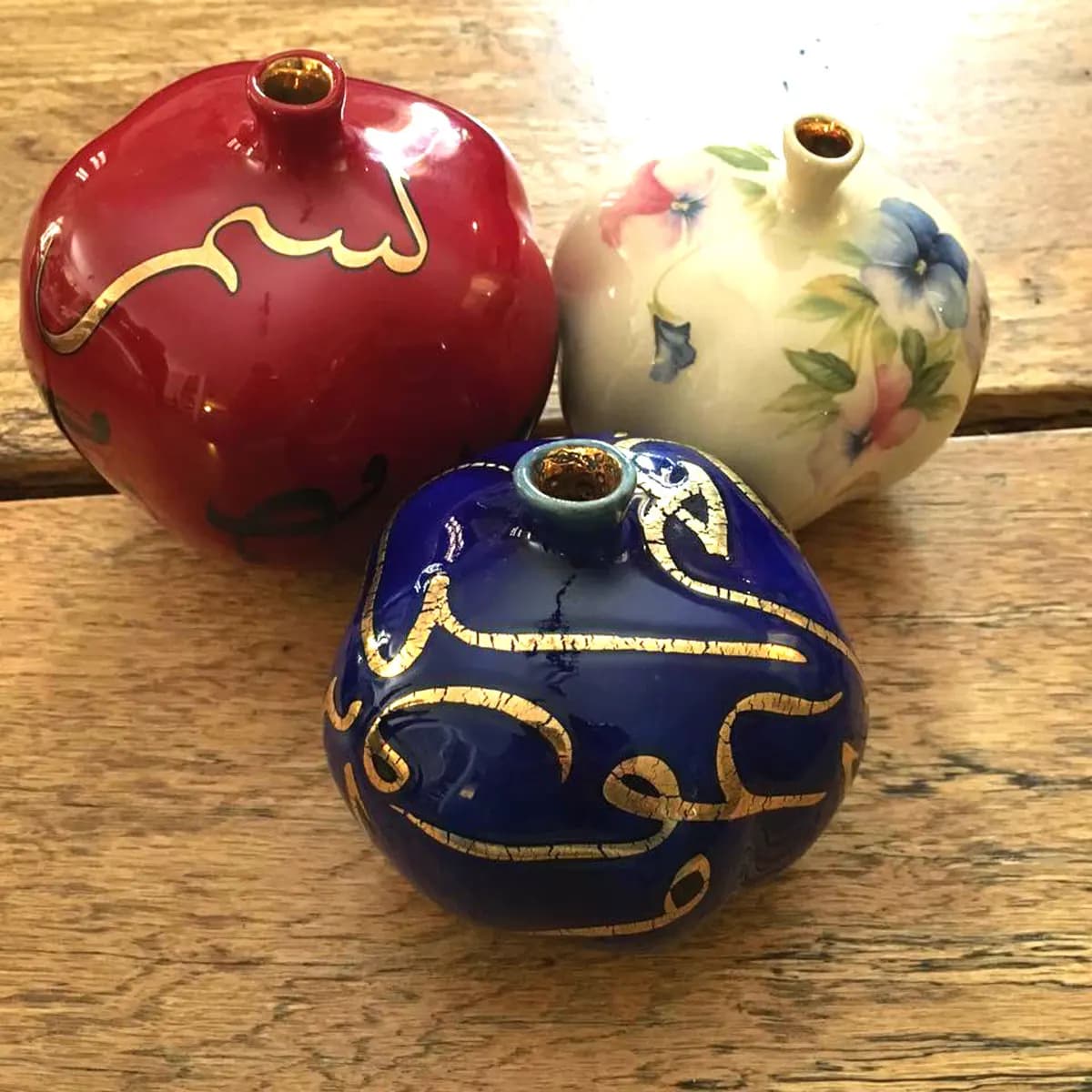 Three glazed ceramic pomegranates in crimson, cobalt and ivory-floral finishes, each rimmed in gold